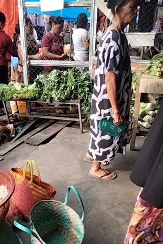 The video shows a market scene with several people. In the foreground, an elderly woman wearing a black and white patterned dress and flip-flops stands holding a green purse. Behind her, a person wearing a blue shirt and jeans is visible. To the right, a large wooden crate is filled with shallots. In the background, other individuals are seen interacting at stalls, with various goods and produce displayed. The environment appears to be an outdoor or semi-outdoor market with a concrete floor and some fencing. The footage is oriented vertically, with the action appearing to move from the bottom to the top of the frame.