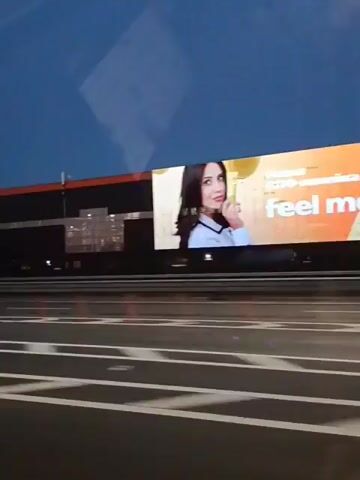 A nighttime exterior shot shows a large, dark-colored building with orange trim along the top. The building appears to be a warehouse or industrial facility. In the foreground, a highway with white lane markings is visible. The camera is positioned inside a vehicle, likely a car, as a side mirror is partially visible in the lower left corner. A bright billboard with an image of a woman is displayed on the side of the building. Reflections of lights and the sky are visible on the windows of the building. The video appears to be filmed while moving, as the perspective shifts slightly.