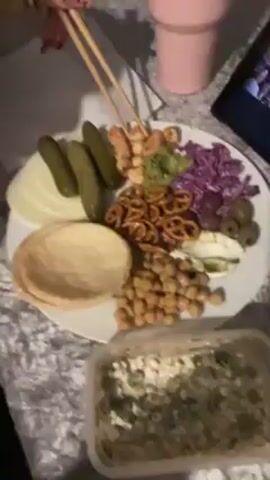 A woman eats something with chopsticks. She holds up a pale food item and puts it into her mouth, and then returns the camera to a plate of snacks and appetizer-like food. The plate contains chickpeas, pretzels, pickles, sliced onions, a pile of what appears to be red cabbage, some flat pita-like bread, and some olives. In the bottom of the frame there is also a partially visible container holding a salad of some kind. The video appears to be shot in a low-lit indoor environment.