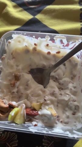 A close-up shot focuses on a clear plastic container holding a creamy dessert. A spoon is inserted into the mixture, which appears to contain nuts, fruits, and a thick, white sauce, possibly yogurt or cream. The dessert is on a yellow and black patterned surface. The scene is brightly lit.