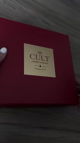 The video begins with a close-up of a closed, dark red gift box with a gold label that reads "Be CULT CHERRY ESSENCE FRAGRANCE GIFT SET". A person's hands with white fingernail polish are holding the box. The box is then opened to reveal its contents: a dark red perfume bottle with a gold cap, a red tube of lotion with a white cap, and the interior of the box is decorated with a pink pattern featuring cherries. The camera pans slightly as the box is opened.
