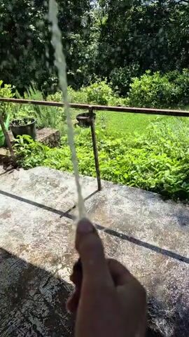 A person's hand holds a black object, possibly a hose nozzle, from which a stream of water is being sprayed. The water is clear and appears to be under pressure, creating a fine mist and droplets. The background shows lush green foliage and trees, suggesting an outdoor, possibly garden or yard setting. The ground in the foreground is made of concrete and appears wet, with a shadow cast upon it. A metal railing runs horizontally across the middle ground. The scene is lit by natural sunlight, creating bright conditions.