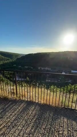 A vertical video displays a scenic view of a valley at sunset. The foreground is dominated by a rustic metal fence and a rocky, grassy ledge. Beyond the fence, a winding road snakes through a lush green valley dotted with trees and a few buildings. The sun is setting on the right side of the frame, casting a warm glow over the landscape and creating long shadows. The sky is clear and a vibrant blue, transitioning to a lighter hue near the horizon. The video captures the tranquil beauty of nature as day turns to night.