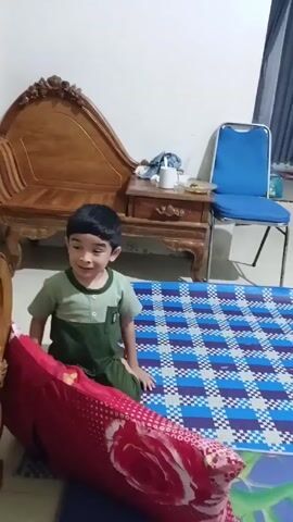 A young boy is seen on a checkered mat, leaning forward and then lying down. A young girl is seen eating something that resembles a small bun. She then reaches her hand out toward the camera. The scene takes place inside a room that includes an ornate wooden settee, a small side table, and a blue chair. A red pillow with flowers on it is also visible. The walls are white.