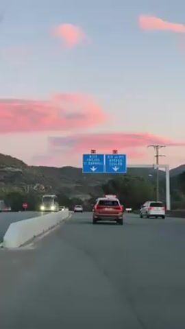 The video depicts a highway scene at dusk. Several vehicles, including a bus and cars, are traveling on the road. Above the lanes, blue signs point directions to Nice, Fréjus, St. Raphael, Aix-en-Provence, Hyères, and Toulon. The sky is pale with pink-tinged clouds. Mountains are visible in the background. The overall impression is of a typical road trip scene during sunset.