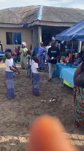 Five children, three girls and two boys, dance on a dirt patch, clapping their hands and raising their arms. They are dressed in white shirts and long, blue and white patterned skirts. An adult man in a black t-shirt and light blue jeans stands nearby, holding papers and observing. Several adult women, some in white head coverings and long dresses, watch from the background. A building with a blue roof is visible in the background, along with a blue canopy and tables. A person's hand with an orange fingernail is visible in the foreground.