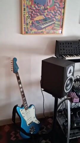 The video shows a small music studio. There's a blue electric guitar leaning against a wall, with two framed artworks hanging above it. A black keyboard synthesizer sits on a stand. In front of the keyboard, there's a black monitor speaker mounted on a stand. A metal rack holds various audio equipment including pedals and other controllers. The studio contains numerous keyboards, controllers, and other devices. A microphone stand with headphones attached stands near a window. The room has white walls and a carpet.