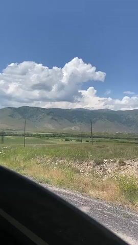 A time-lapse video shows a passing view from a moving vehicle. The scene is a rural landscape with rolling green hills and mountains in the background under a bright blue sky with scattered white clouds. In the foreground, there is a dirt road or path with grass and wildflowers growing alongside it. There are also power lines visible in the distance. The camera captures the passing of time, with the light and shadows subtly shifting, suggesting movement through the landscape.