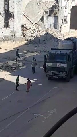 A military green truck with the MSF logo is parked on a street. The truck is loaded with large white water tanks. A group of people, including children, are gathered around the back of the truck. Some individuals are reaching into the truck bed, seemingly to unload or distribute supplies. Other people are standing nearby, some carrying empty containers. In the background, there are damaged buildings, suggesting a war-torn or disaster-affected area. The ground is paved and appears somewhat dusty. The lighting indicates it is daytime.