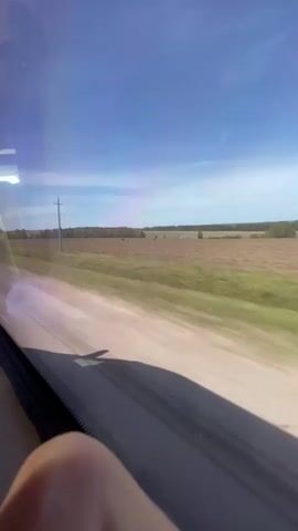 The video shows the view from a moving vehicle, likely a bus or car, with a field visible through the window. A clear blue sky with some thin clouds is seen above the landscape. The field appears to be mostly brown with patches of green, possibly agricultural land. There are power lines and poles along the edge of the field. The shadow of something inside the vehicle is cast on the lower portion of the window. The video is slightly shaky, indicating movement.