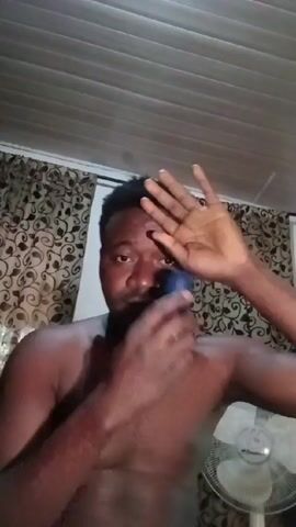 A shirtless man applies roll-on deodorant to his armpit, which he then rubs in. The man has dark skin, dark hair, and a slight mustache. He is wearing a thin necklace. The environment features a white, horizontally-slatted ceiling, a window with a paisley curtain, and a white electric fan with silver accents.