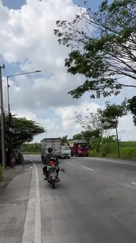 The video shows a roadside scene with a paved road and a sidewalk. On the road, a motorcycle with two people is traveling away from the camera. Behind the motorcycle, a truck is also moving in the same direction. Further down the road, a white truck is approaching the camera. The environment is a rural or semi-rural area with trees and vegetation on the sides of the road. The sky is partly cloudy with white clouds. There is a lamppost on the left side of the road, and some structures are visible on the sidewalk. The overall impression is of a typical day on a road with traffic.