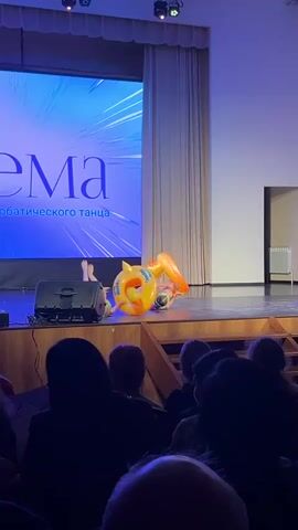 The video shows a performance on a stage in a dimly lit hall, with an audience in the foreground.  On the stage, two inflatable yellow ducks with rider seat designs rest on a brown-wooden floor, one on each side of a silver speaker. Between the ducks, two legs extend upwards, presumably from a performer doing a handstand. A large screen behind the stage displays an illuminated blue background with the text "СЕМА" in white and smaller text underneath. The curtains behind the stage are closed. The audience is silhouetted. The overall atmosphere suggests a dance performance or show. The video is shot from a static position at a distance from the stage.
