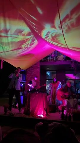 A band is performing on a stage outdoors. The stage is covered by a large, draped canopy with vibrant pink and yellow projections of abstract shapes and lines. The canopy is lit from above with pink and yellow lights. The band consists of at least four members. On the left, a man in a light-colored jacket and dark pants is playing a trumpet. In the center, a person is standing behind a table covered with a sequined pink cloth, with equipment that appears to be for DJing or electronic music production. Further to the right, a person wearing a brightly colored, patterned robe is standing and possibly singing or playing an instrument. To the far right, a person is seated, playing an acoustic guitar. The background shows a building with dark windows and a balcony. The foreground is dark, suggesting an audience is present but mostly out of frame. The overall atmosphere is colorful and festive.