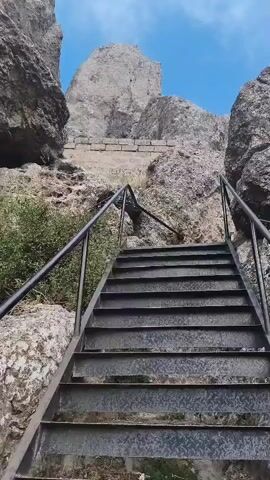 The video shows a set of outdoor metal stairs with black railings ascending a rocky incline. The stairs appear to be made of diamond plate metal. To the left of the stairs, there is dense green shrubbery. Large rock formations dominate the landscape, with a rough-hewn stone wall or structure visible further up the incline. The sky above is a clear blue with a few wispy clouds. The camera perspective is looking up the stairs, suggesting movement or an intended ascent. There are no people visible in the video.