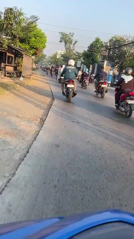 A POV shot from a blue motorcycle shows a line of motorcycles riding down a paved road. The motorcycles are mostly in a single file line, with some scattered on either side of the road. The road is lined with trees and houses on the left, and buildings and more trees on the right. The sky is clear and the sun is shining. The motorcycles appear to be heading towards a busy intersection with other vehicles, including cars and trucks. Some of the motorcyclists are wearing helmets, while others are not. The overall impression is of a busy street scene in a tropical or subtropical region.