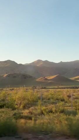 The video shows a landscape featuring rolling hills and a clear blue sky. The foreground is a field of tall grass and sparse vegetation, with a fence running parallel to the camera's movement. As the camera moves, trees are briefly visible. The hills in the background are bathed in sunlight, creating shadows and highlights that emphasize their contours.