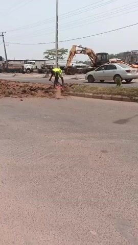 The video shows a man working on a road construction site. He is wearing a neon yellow vest and is using a shovel to clear dirt and debris from the road. In the background, there is a silver car parked on the side of the road, and an excavator and other construction vehicles.  A telephone pole is in the background. The road is a paved surface. The weather appears to be overcast.
