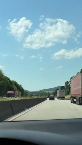 The video is filmed from the interior of a moving vehicle, looking out through the windshield. It shows a highway scene on a clear day with blue skies and scattered white clouds. The highway has multiple lanes, and traffic includes several vehicles, primarily trucks and cars. On the left side of the highway, there is a concrete barrier, followed by trees and green landscape with rolling hills in the distance. On the right side, there are trees and more open landscape. The camera perspective suggests the vehicle is in the right lane, with a large red and white truck visible in the foreground on the right. The overall environment appears to be rural or semi-rural.