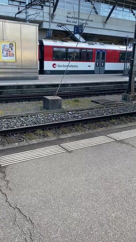 The video shows a train at a train station. The train is white with black and red stripes, and the words "SBB CFF FFS" are printed on the side. The train is pulling away from the platform, and the camera follows it as it moves. The station is covered by a metal roof, and there are advertisements posted on the walls. The ground is paved with asphalt and marked with white safety lines.