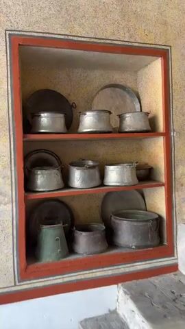 The video shows the interior of a building, likely a historical or religious site. A built-in wooden shelf painted with red trim is filled with old metal cooking pots and platters of various sizes and finishes. To the right of the shelf is a concrete structure that appears to be a water fountain or basin. Above the basin are decorative arches painted with remnants of green and red. An oval plaque with a carved design is centered above an inscription written in a Cyrillic-based script. A metal ladle hangs from one of the faucets. The overall aesthetic suggests a rustic and historical environment.