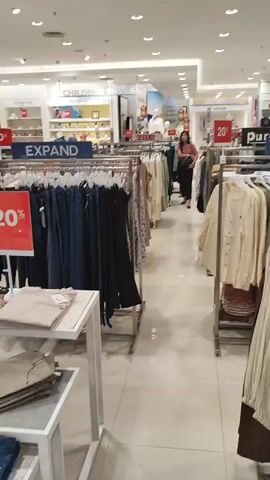 The video shows the interior of a clothing store. Racks of women's clothing are visible on both sides of a walkway. The clothing consists of dresses, pants, and tops in various colors and styles. A woman in a pink shirt and black pants walks down the aisle in the middle of the store, appearing to browse the merchandise. Signs indicating a 20% discount are displayed. The store is well-lit with fluorescent lights and has a clean, organized appearance.