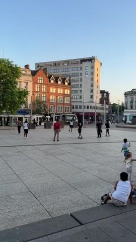 The video shows a group of young men playing keep-up with a soccer ball in a public square. The square is paved with large, light-colored tiles, and is surrounded by buildings of varying heights and architectural styles. There are shops like Costa Coffee and Starbucks, as well as a tall building with the word "PEARL" on it. The sky is clear and blue, indicating a sunny day. There are several people walking around the square, and a few children sitting on a low wall. The camera follows the ball as the young men kick it to each other, aiming to keep it in the air. The camera appears to be held by someone standing at the edge of the square.