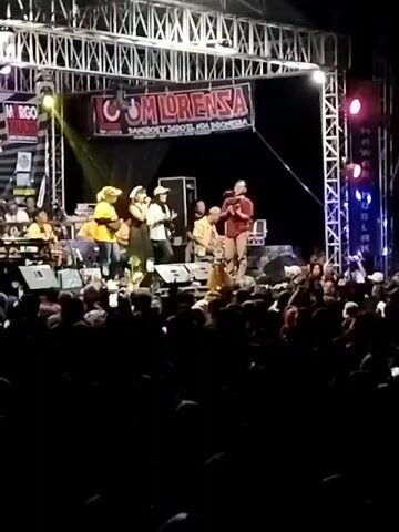 A band called "GUM LORENSA" is performing on an outdoor stage at night, in front of a large audience. The stage is set up with a large banner that reads "GUM LORENSA" and a smaller banner below it that is unreadable. There are several band members on stage, including vocalists and musicians playing instruments. The audience is packed together in front of the stage, with many people holding up their phones to record the performance. The lighting is dramatic, with spotlights illuminating the stage and creating a colorful atmosphere. The overall scene suggests a lively and energetic concert event.