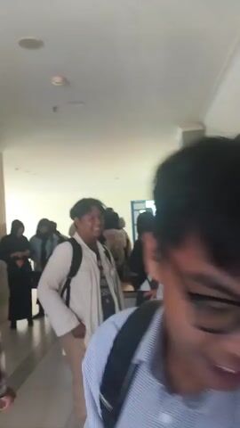 A group of students are walking down a hallway. Some are wearing backpacks. One person in the foreground is wearing a white jacket and tan pants and appears to be talking and smiling. In the background, several other students are visible, some wearing hijabs. An elevator is visible on the right side of the frame. The hallway appears to be well-lit with fluorescent lighting.