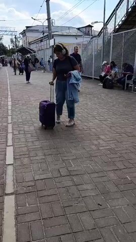 The video shows a train station platform. In the foreground, a woman with curly brown hair, wearing headphones and a denim jacket over a dark t-shirt, stands with a purple suitcase on wheels. She is looking at the camera. Behind her, a man walks away from the camera. Further down the platform, other people are visible, some sitting on benches and others walking. A fence runs along the edge of the platform. A small red building, possibly a kiosk or ticket booth, is visible behind the fence. The ground is paved with bricks. The sky is overcast.