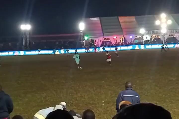 The video depicts a soccer game at night, with floodlights illuminating the field. Players in red and green jerseys are visible on the grass, some running. The view is from behind spectators, whose heads and backs are visible in the foreground. A large tent-like structure is in the background, decorated with lights. An LED screen runs along the side of the field. The atmosphere seems lively, potentially a well-attended event.