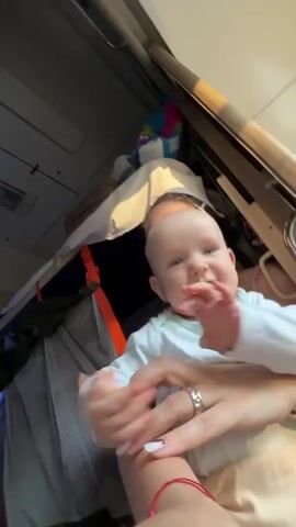 A baby is held by a person whose face is partially visible behind the baby. The baby is wearing a white long-sleeved shirt and light-colored pants. The baby's hands are near its mouth. The person holding the baby has tattoos visible on their arm. They are in what appears to be a train compartment, with luggage visible in the background. The lighting suggests artificial light.
