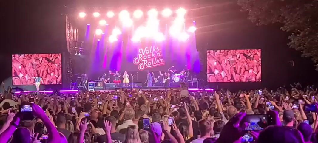A concert is in progress with a band on a brightly lit stage performing in front of a large crowd of people. The stage is decorated with the words "Volks Rock'n Roller" in a stylized font. Two large screens on either side of the stage display close-ups of performers. The crowd is holding up many cell phones to record the event. The lighting is predominantly purple and white.