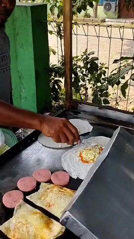 A person is preparing food on a flat griddle. There are several round, pink slices of what appear to be sausage on the left side of the griddle. In the center, two white tortillas are being filled with ingredients that include shredded cabbage, a creamy white substance, and an orange sauce. The person is using a small, silver utensil to spread the filling. Several portions of folded, cooked tortillas are visible on the lower part of the griddle. The griddle is placed outdoors in front of a green wall and a metal fence. The background shows some foliage and a building with a window. The lighting suggests it is daytime.