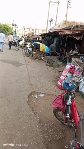 The camera pans along a street lined with small, open-front shops and parked motorcycles. Several people walk and stand near the shops, some are sitting. One person sits behind a table inside a shop. A cart loaded with clothing sits on the edge of the road. The road has a pothole and some trash. Boxes are stacked beside a red scooter. Overhead power lines are visible. The video is shot during the day.