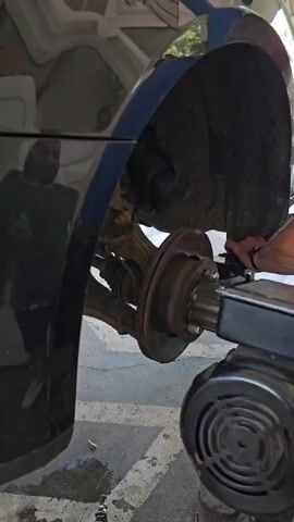 The media file shows a close-up, low-angle shot of a car's brake rotor being machined. The car is black, and the wheel well is visible, revealing the brake assembly. A person's hand, wearing a dark glove, is seen operating a tool that is attached to the rotor. The tool appears to be a brake lathe, a device used to resurface brake rotors. The setting appears to be an outdoor area, possibly a driveway or garage, with a concrete floor and some debris scattered around. The lighting suggests it is daytime, but no specific time or weather conditions are evident. There is no visible text in the media file. The overall mood is focused and practical, suggesting a repair or maintenance activity. The reflection of a person in the car's bodywork is also visible, but their features are indistinct.