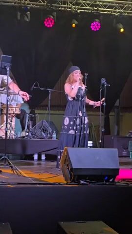 This is a video of a live music performance on a stage. In the foreground, two singers are visible. The male singer on the left is wearing a light-colored short-sleeved shirt and gray pants, and is holding a microphone and looking at a tablet on a stand. The female singer on the right is wearing a black dress with a celestial pattern and a headscarf, and is also holding a microphone. Behind them, a band is playing, including a drummer on the right and a guitarist on the left. The stage is lit with colored lights, including pink, blue, and yellow. There are speakers and other audio equipment on the stage. The background appears to be an outdoor venue with a large structure behind the performers. The video is filmed from the audience's perspective.