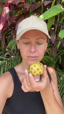 A woman in a black tank top and a tan baseball cap with the word "INCENSE" printed on it holds a yellow, bumpy fruit up to her nose, inhales, and then grimaces, closing her eyes and opening her mouth as though something smells unpleasant. She holds the fruit in her right hand. Behind her are red, pink, and green plants.