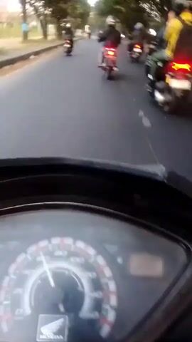 A first-person perspective video shows a group of motorcyclists riding down a paved road lined with trees. The camera appears to be mounted on the handlebars of a scooter or motorcycle, focusing on the instrument panel in the foreground. The speedometer is visible, with the needle indicating a speed of approximately 30 kilometers per hour. In the background, several other motorcycles are visible, some with riders wearing helmets. The overall scene suggests a casual group ride on a clear day.