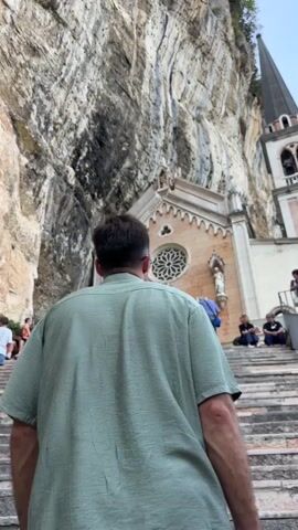 A man wearing a light green short-sleeved shirt and white pants walks up a wide stone staircase towards a church built into a cliff face. The church has a pale pink facade with arched windows and a pointed spire. Several people are seated on the stairs on the left side of the frame, and a few more are visible on the right, near the church entrance. The cliff face behind the church is large and rocky, with a lighter colored surface. The sky above is hazy. The man is viewed from behind, and he is carrying a white bag in his right hand. He is nearing the top of the stairs.