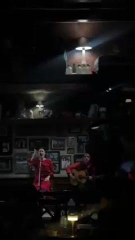 A video shows a musical performance in a dimly lit venue. A woman in a red patterned outfit is singing into a microphone, gesturing with her hands. To her right, a man is playing an acoustic guitar. They are on a small stage in front of an audience. Behind them is a wall lined with framed pictures and shelves with various objects, illuminated by small lamps. A few members of the audience are visible in the foreground, one with a lit candle on their table.