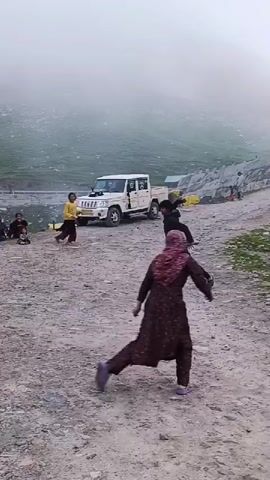 A group of people are gathered in a mountainous area with fog. A white pickup truck is parked on the side of the road. Some individuals are playing a game, running and interacting with each other on a gravel and dirt path. In the background, there are green hills and more fog.