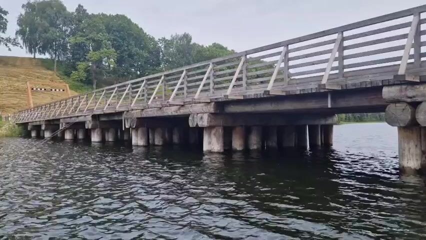 A long, weathered wooden bridge with a railing extends over dark water. The bridge is supported by numerous vertical wooden pilings. On the left bank, a grassy slope rises, with some trees and a fence visible. The sky is overcast. The camera is positioned low, near the water's surface, and is moving slowly to the right, giving a slightly unsteady view of the bridge. There are no people or animals visible in the frame. The overall impression is one of a rustic, somewhat aged structure in a natural setting.