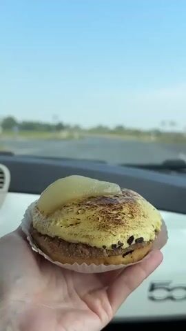 A hand holds a pastry in front of the windshield of a car. The pastry has a golden-brown toasted top with a slice of fruit on top. The car is driving on a road through a rural area with fields and trees on either side. The sky is blue with a few clouds.