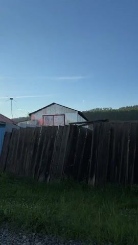 The video shows an outdoor scene with a wooden fence in the foreground. Beyond the fence, there are several old, rustic wooden buildings, including a barn or shed with a corrugated roof and what appears to be a log cabin. The buildings are surrounded by tall green grass and vegetation. In the background, there are rolling hills covered in trees. The sky is a clear blue with a few wispy clouds. The lighting suggests it is daytime, likely late afternoon or early morning due to the low angle of the sun casting shadows. There are no people visible in the video. The overall impression is one of a rural, somewhat neglected setting.