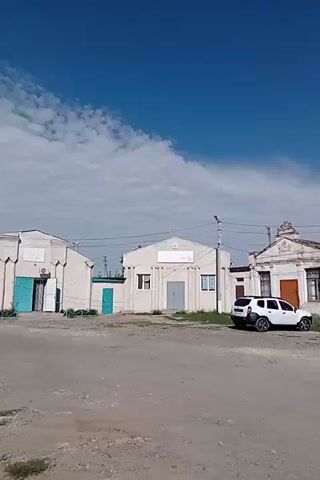 The video pans across a street in a rural or small-town setting. The sky is bright blue with scattered clouds. The buildings lining the street appear to be a mix of older, somewhat dilapidated structures and newer, more modern ones.

On the left side of the street, there's a long, light-colored building with a large, flat facade and a darker, possibly metal roof. A white SUV is parked in front of it. Further down, there's another building with a more decorative facade, featuring arched windows and ornate detailing. A blue sedan is parked in front of this building.

In the center of the frame, there are several smaller buildings, some with colorful paint and shop signs. A white car is parked in front of one of these. On the right side, there are more buildings, including one with a light pink facade. A blue car is visible further down this side of the street.

In the foreground, the ground appears to be unpaved, with dirt and some patches of grass. There are also some concrete structures, possibly remnants of a larger construction or infrastructure, visible towards the right side of the frame. The overall impression is of a quiet street scene, possibly in Eastern Europe or a similar region, with a blend of old and new architecture.