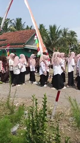 A group of young people in uniforms, some in brown scout-like attire and others in white shirts and headscarves, are walking in a procession or parade. They are outdoors on a paved surface, possibly a road or path, with a building with a red tiled roof visible in the background. Several Indonesian flags, red and white, are displayed prominently, some flying from poles and others attached to a tall, diagonal structure. The weather appears to be sunny. The individuals are mostly facing towards the right of the frame, suggesting they are moving in that direction. Some individuals are holding sticks or poles. Lush green vegetation is present in the foreground.
