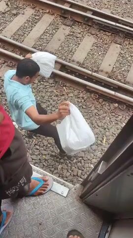The video shows a train station or railway track environment. In the foreground, a person's leg is visible wearing a blue flip-flop, standing on a metal platform of what appears to be a train door. Beside them, another person is holding a white plastic bag. The camera pans to show the railway tracks stretching into the distance. Several sets of tracks are visible, covered in gravel and ballast. In the background, a small white building with a sign, possibly a signal box or station office, is present. Several people are standing near this building, some appearing to be railway workers. A tall water tower is also visible in the background, along with trees and overhead power lines for the train. The overall atmosphere is that of an active railway location.