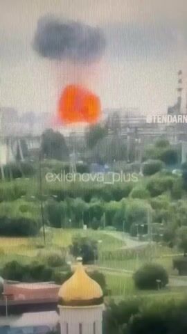 A video shows a large plume of smoke rising into the sky, with a bright orange glow at its base, indicating a fire or explosion. The smoke is dark gray and billows upwards, expanding as it dissipates. Below the smoke plume, buildings and vegetation are visible, suggesting an urban or industrial setting. The image quality is poor, with a visible grid pattern and low resolution, making it difficult to discern fine details. Watermarks in the video read "exilenova_plus" and "@TENDARN".