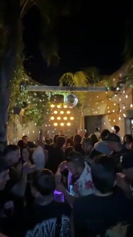 A crowd of people are gathered in what appears to be an outdoor nightclub or dance venue. The lighting is dark, with a disco ball hanging from above, casting spots of light across the walls and people. Some people are wearing casual clothes like t-shirts and caps, while one individual in the foreground is wearing a floral shirt. The setting appears to be an enclosed outdoor space, with walls and possible plant growth adding to the ambiance. Some people are holding up their phones, possibly recording or taking pictures. A person in the foreground is opening a bottle. The overall atmosphere is lively and social.